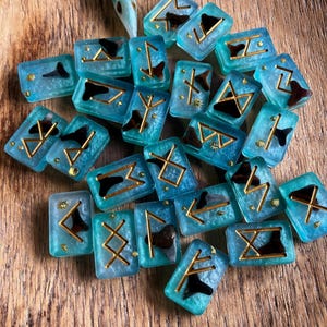 May include: A collection of rectangular resin tiles in a translucent teal color, each embedded with a dark shark tooth and a gold-colored rune symbol. A small, light blue bird skull is positioned at the top of the scattered arrangement.