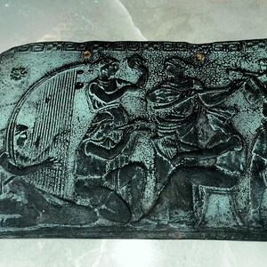 May include: A dark green rectangular plaque with a raised relief design depicting a group of people in a scene. The plaque has a decorative border.