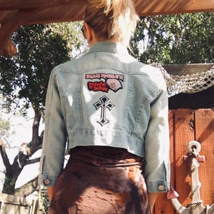May include: A light blue denim jacket with a cropped hem. The jacket has a black cross and a patch with the text "Iron Maiden" and an image of Eddie, the band's mascot.