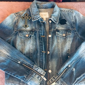 May include: A blue denim jacket with a black cross and a black patch on the front. The jacket has zippered cuffs and a button closure.