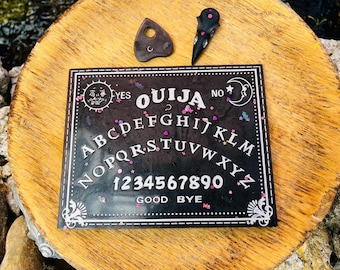 Könnte beinhalten: Ein schwarzes, rechteckiges Ouija-Brett mit weißen Buchstaben, Zahlen und Himmelszeichen, das mit buntem Glitzer versehen ist. Über dem Brett liegen eine dunkelbraune, herzförmige Planchette und ein schwarzer Zeiger in Form eines Vogelschädels.