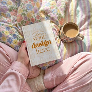 May include: A book with the text "YOUR design HERE" is held in a person's hand. The book has a white cover with a basket pattern. A floral mug with coffee sits nearby on a bed with floral and striped bedding. The person is wearing pink pajamas.