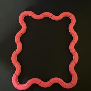 May include: A coral-colored, wavy-edged, square-shaped toy. The toy is made of connected segments, forming a flexible, geometric shape. The toy is set against a black background.