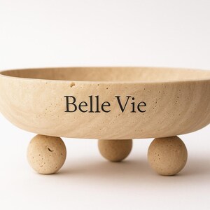Op de afbeelding: Een beige, ronde kom met drie bolvormige voetjes. De woorden "Belle Vie" staan op de zijkant gedrukt. De kom is gemaakt van een lichtgekleurd, getextureerd materiaal en staat op een witte achtergrond.