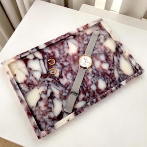 Op de afbeelding: Een rechthoekige tray met een marmerpatroon in tinten bordeauxrood en wit. De tray bevat een goudkleurig horloge met een zilveren band en twee gouden oorringen. De items zijn gerangschikt op een wit oppervlak.