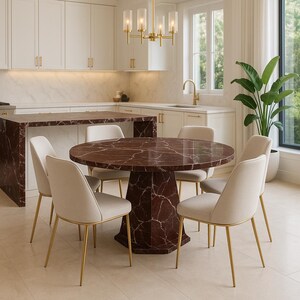 Op de afbeelding: Een moderne eethoek met een ronde tafel van donkerrood marmer met een geometrische voet. Zes crèmekleurige stoelen met gouden poten omringen de tafel. Een gouden kroonluchter hangt erboven.