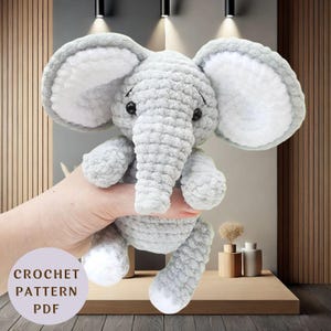 May include: A handmade grey crochet elephant toy with large ears and a trunk. The elephant has white accents on its ears and feet. The text "CROCHET PATTERN PDF" is in the lower left corner. The elephant is a soft toy.