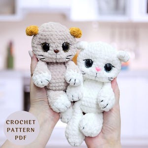 Può includere: Due giocattoli a forma di gatto all'uncinetto, uno beige con orecchie gialle e uno bianco. Entrambi hanno occhi ricamati e nasi rosa.