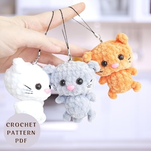 Peut inclure: Trois porte-clés en forme de chat crochetés en blanc, gris et orange. Les porte-clés sont suspendus à une main. L'image comprend le texte "CROCHET PATTERN PDF".
