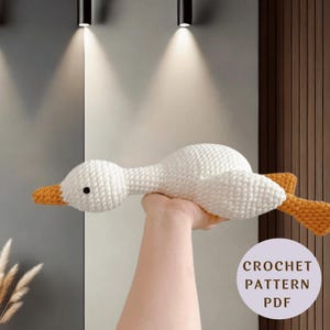 Può includere: Un giocattolo di oca all'uncinetto bianco con becco e zampe arancioni. L'oca ha un occhio nero ed è realizzata in un materiale a maglia strutturato. L'immagine include una grafica circolare che dice "CROCHET PATTERN PDF".