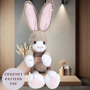 May include: A handmade crochet bunny plushie with long ears, a white muzzle, and pink accents on the ears and feet. The bunny is brown and white, with black eyes and a pink nose. The text "CROCHET PATTERN PDF" is visible.