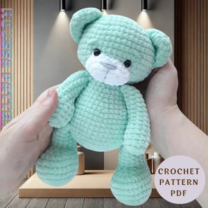 May include: A crocheted mint green teddy bear with white facial features. The bear is being held by a person's hands. The text "CROCHET PATTERN PDF" is in the bottom right corner of the image.