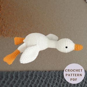 Puede incluir: Un ganso de ganchillo blanco con patas y pico de color naranja, que presenta un pequeño ojo negro. Una insignia circular de color morado claro en la esquina inferior derecha contiene el texto CROCHET PATTERN PDF en letras mayúsculas marrones.