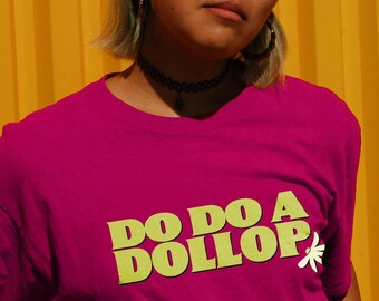 Może przedstawiać: Osoba w różowej koszulce z napisem "Do Do A Dollop" w żółtych literach. Osoba nosi okulary okrągłe i ma włosy związane w dwa kucyki.