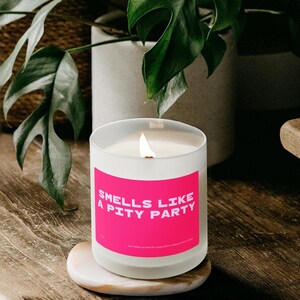 Peut inclure: Une bougie blanche allumée dans un pot en verre transparent avec une étiquette rose vif qui dit "Smells Like a Pity Party". La bougie repose sur un petit dessous de verre rond en pierre de couleur claire. Une deuxième bougie avec un graphique de surfeur est visible.