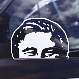 May include: A black and white sticker of a person's face with a serious expression. The sticker is on a car window.