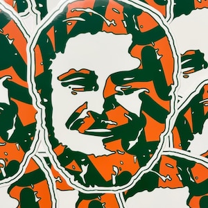 May include: A sticker with a white background and a green and orange design. The design is a stylized portrait of a person with a mustache.