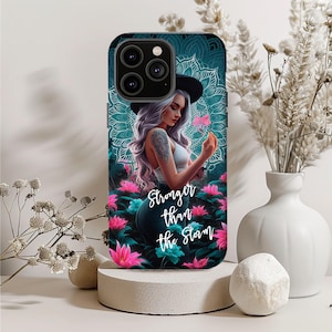 May include: A phone case with a teal background, a woman with purple hair, a hat, and a tattoo, holding a flower. The case has the text "Stronger than the Storm" and a floral design.