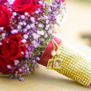 May include: A bridal bouquet with red roses, purple orchids, and white baby's breath. The bouquet is wrapped in a gold fabric with a beaded trim and has two small framed photos attached.