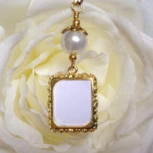 May include: A gold-toned charm with a white pearl and a square frame with a white background. The charm is hanging from a white rose.
