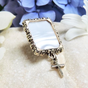 A silver tone lapel pin featuring a small, floral design picture frame with a tiny cross  dangling from it. A lovely way to honour  someone you love.