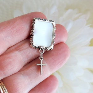 A silver tone lapel pin featuring a small, floral design picture frame with a tiny cross  dangling from it. A lovely way to honour  someone you love.