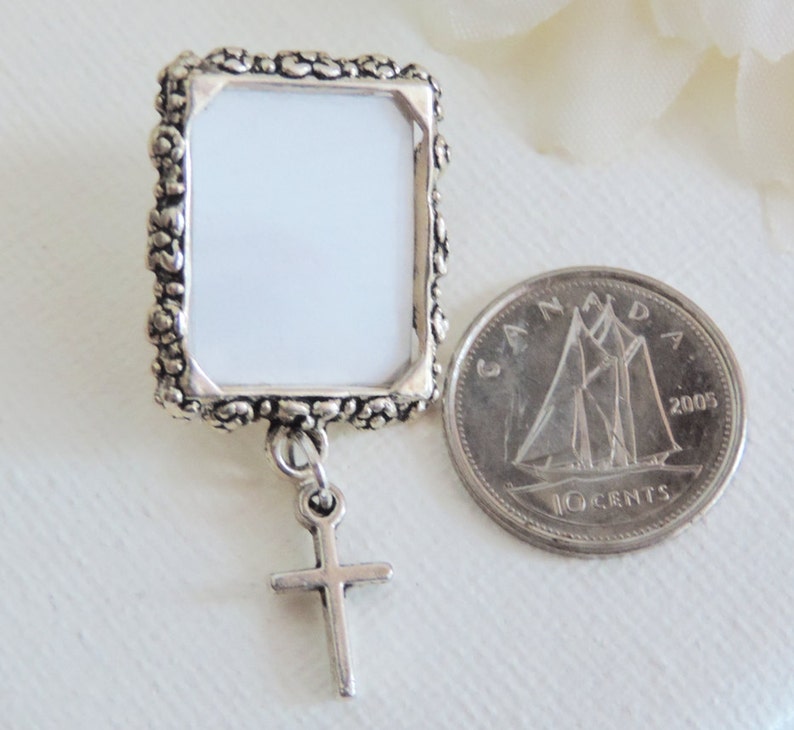 A silver tone lapel pin featuring a small, floral design picture frame with a tiny cross  dangling from it. A lovely way to honour  someone you love.