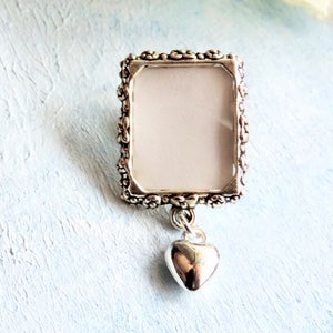 A lapel pin featuring a small, floral design picture frame with a tiny heart dangling from it, all in antique silver tones.