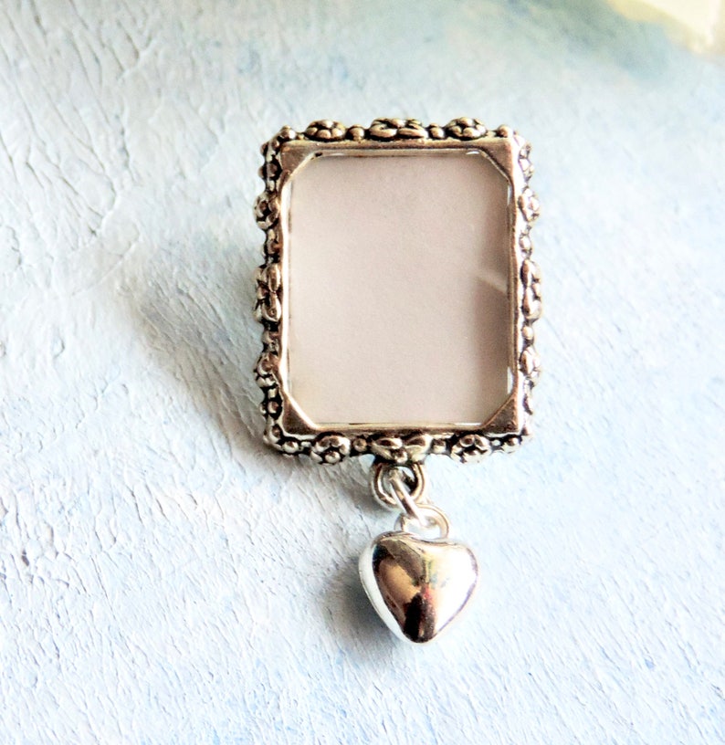 A lapel pin featuring a small, floral design picture frame with a tiny heart dangling from it, all in antique silver tones.