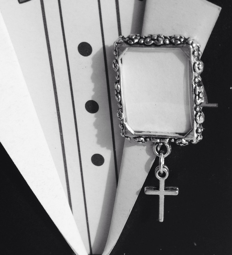 A silver tone lapel pin featuring a small, floral design picture frame with a tiny cross  dangling from it. A lovely way to honour  someone you love.