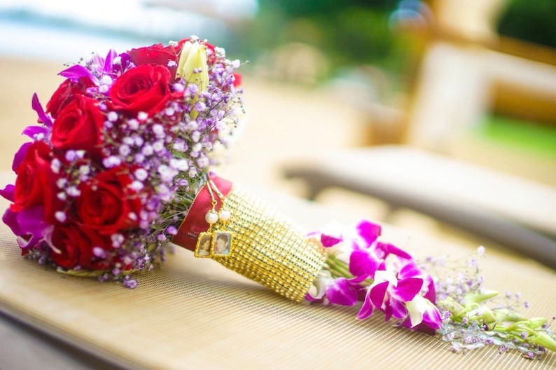 May include: A bridal bouquet with red roses, purple orchids, and white baby's breath. The bouquet is wrapped in a gold fabric with a beaded trim and has two small framed photos attached.