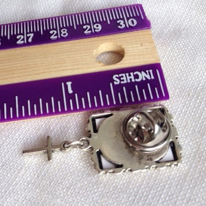 A silver tone lapel pin featuring a small, floral design picture frame with a tiny cross  dangling from it. Shown with a ruler - 1 1/2 inches from top to bottom.