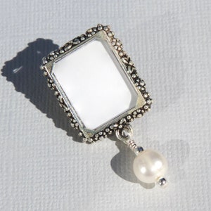 A silver tone lapel pin featuring a small, floral design picture frame with a real freshwater pearl dangling from it.