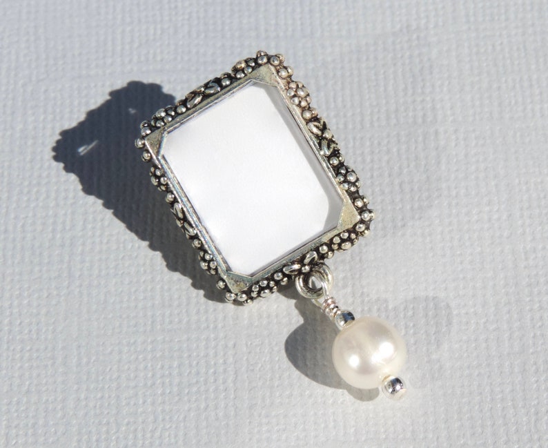 A silver tone lapel pin featuring a small, floral design picture frame with a real freshwater pearl dangling from it.