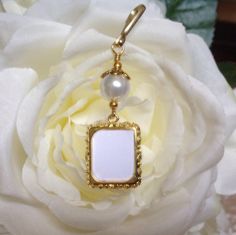 May include: A gold-toned charm with a white pearl and a square frame with a white background. The charm is hanging from a white rose.