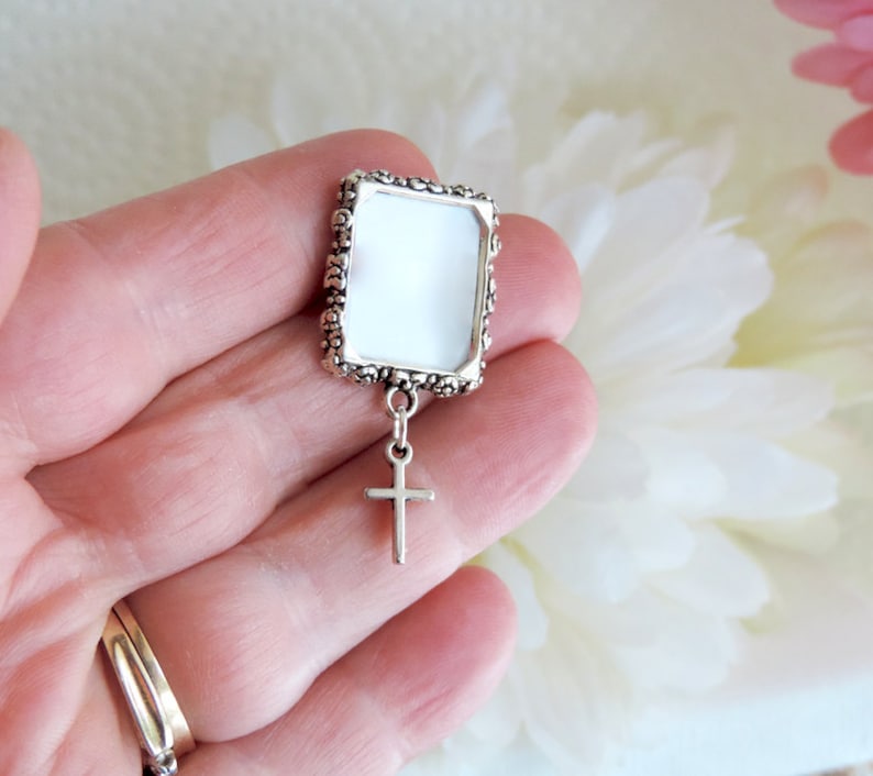 A silver tone lapel pin featuring a small, floral design picture frame with a tiny cross  dangling from it. A lovely way to honour  someone you love.