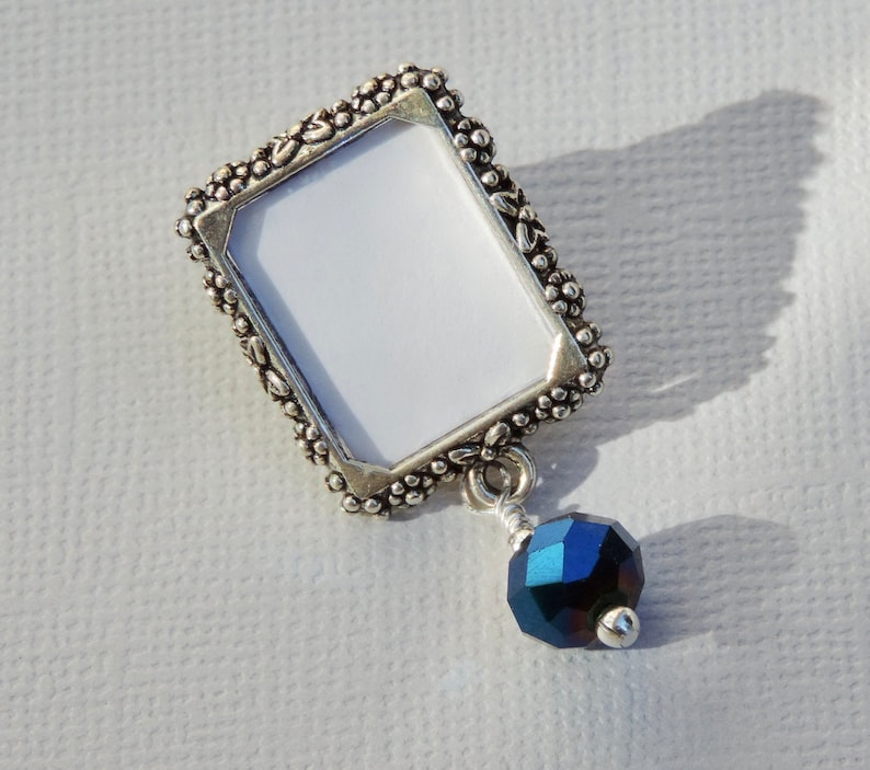 A silver tone lapel pin featuring a small, floral design picture frame with a metallic blue crystal dangling from it.