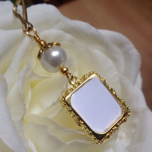 May include: Gold charm with a white pearl and a gold frame with a blank white square inside. The charm is hanging from a gold chain.