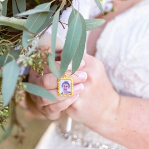 May include: A bride holds a bouquet of white flowers and greenery, with a small gold locket featuring a photo of an older woman attached to the bouquet.