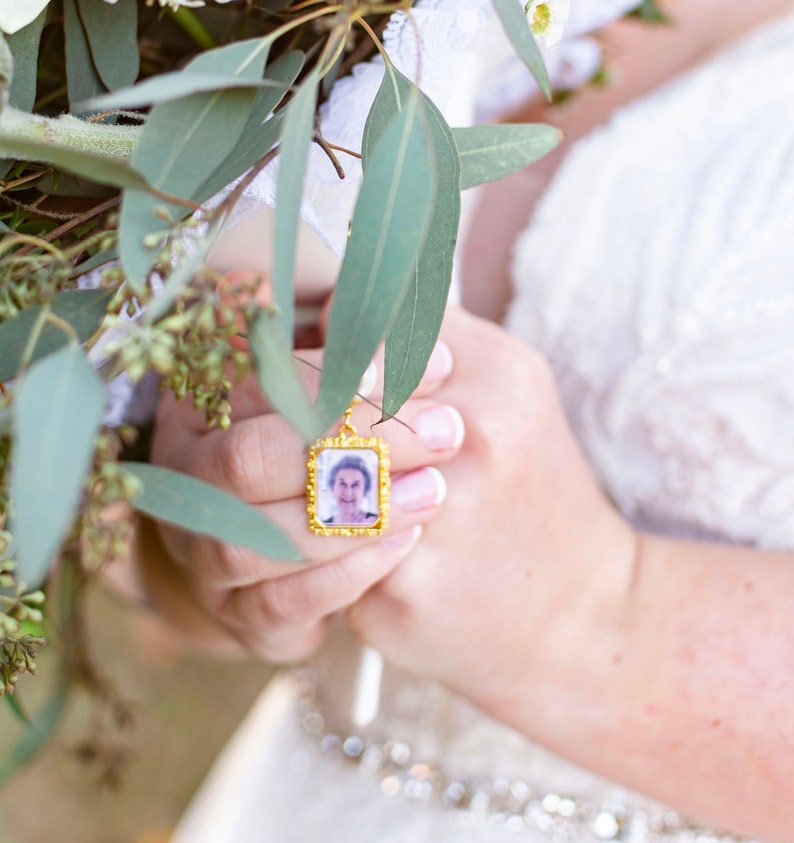May include: A bride holds a bouquet of white flowers and greenery, with a small gold locket featuring a photo of an older woman attached to the bouquet.
