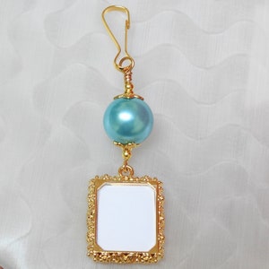May include: Gold picture frame charm with a light blue pearl and a gold clip. The frame is ornate and has a blank space for a photo.