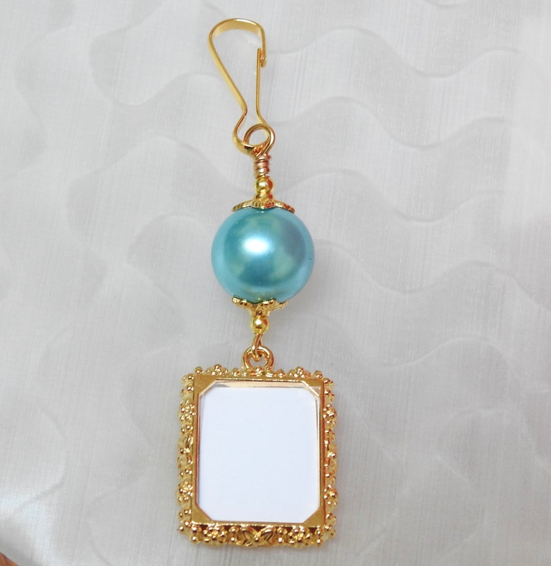 May include: Gold picture frame charm with a light blue pearl and a gold clip. The frame is ornate and has a blank space for a photo.