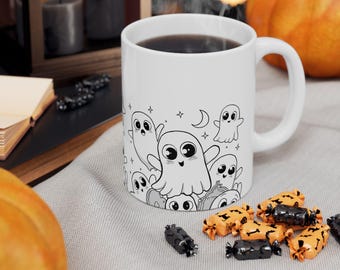 May include: A white ceramic mug featuring a black line art illustration of multiple cartoon ghosts with large eyes, stars, and a crescent moon. The mug is filled with a dark liquid and is surrounded by orange and black wrapped candies.