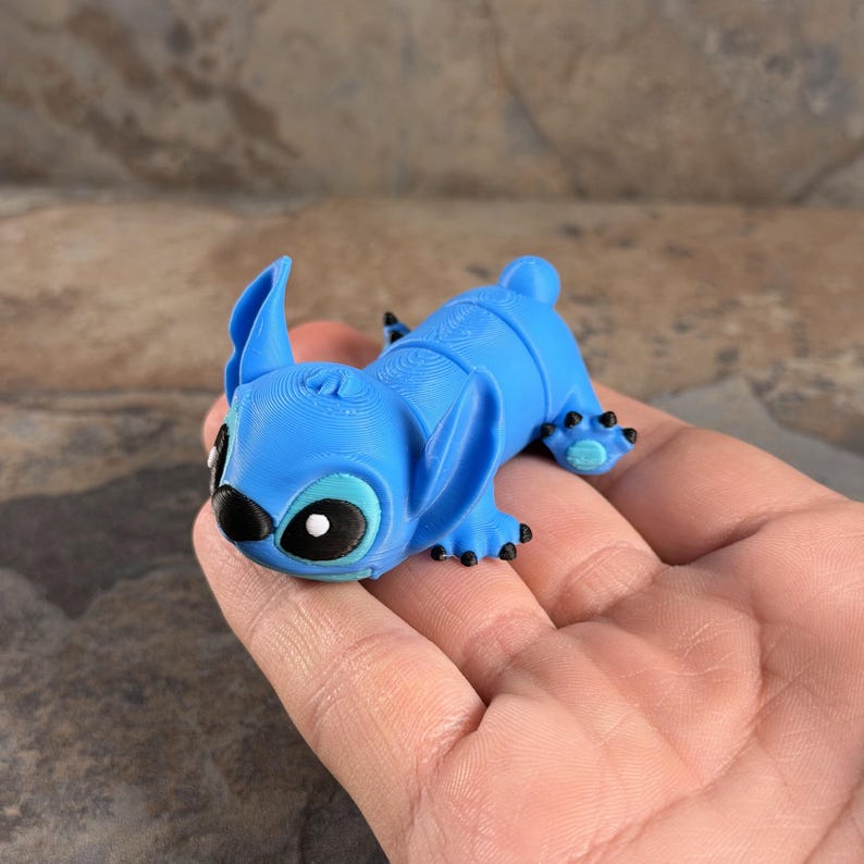 Puede incluir: Una figura azul de un personaje de dibujos animados con orejas grandes, ojos negros y peque&ntilde;as garras negras. La figura est&aacute; tumbada y parece estar hecha de pl&aacute;stico. El personaje es popular.