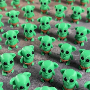 May include: A collection of small, green alien figurines with large ears and black eyes. Each figurine wears a tan diaper. The figures are arranged on a textured, gray surface.