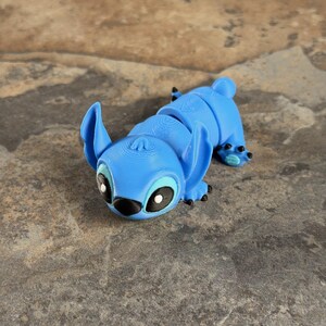 Puede incluir: Una figura articulada azul de un personaje de dibujos animados con orejas grandes y ojos negros. La figura est&aacute; tumbada sobre una superficie texturizada. El cuerpo de la figura est&aacute; segmentado, lo que permite el movimiento. La cara del personaje tiene detalles en blanco y negro.