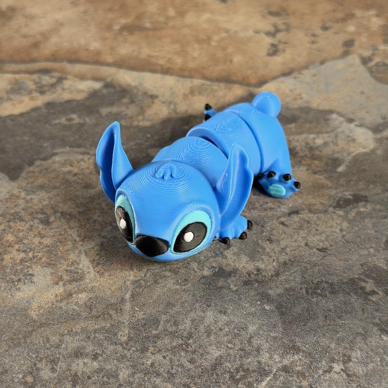 Puede incluir: Una figura articulada azul de un personaje de dibujos animados con orejas grandes y ojos negros. La figura est&aacute; tumbada sobre una superficie texturizada. El cuerpo de la figura est&aacute; segmentado, lo que permite el movimiento. La cara del personaje tiene detalles en blanco y negro.