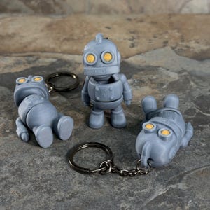 May include: Three gray robot keychains in different poses. The robots have yellow eyes and are attached to silver keyrings. One robot is standing, one is lying down, and one is sitting. The keychains are made of a 3D-printed material.