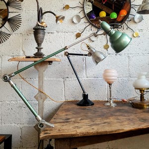 May include: A selection of vintage lamps and a decorative mirror are arranged on a wooden table and against a white brick wall. The lamps include a green articulated desk lamp and a small brass oil lamp.
