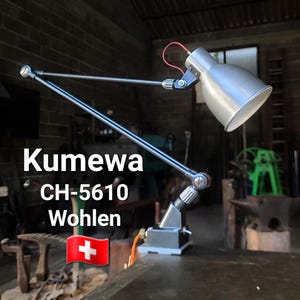 Puede incluir: Lámpara de escritorio plateada con brazo ajustable y pantalla cónica. La lámpara tiene un cable de alimentación rojo y está montada sobre una base rectangular. El texto "Kumewa CH-5610 Wohlen" es visible.